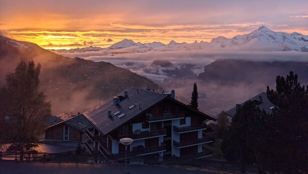 Eine Berghütte mit Balkon und Blick auf schneebedeckte Gipfel und Wolken.