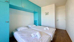 A hotel room with two beds, a teal-colored wardrobe, a small bedside table, and a framed picture on the wall.