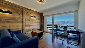 Una sala de estar moderna con una pared decorativa de madera, un sofá, una zona de comedor con mesa y sillas, y un ventanal con vistas a la montaña.