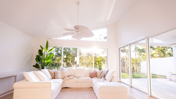 A modern living room with a large L-shaped sectional sofa, a ceiling fan, and large windows offering a view of a garden.