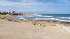 Una playa con costa rocosa, arena y edificios al fondo.