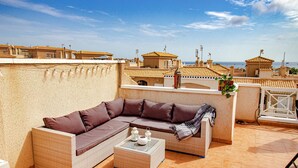 Una terraza en la azotea con un sofá de mimbre, una mesita y vistas al mar y edificios.