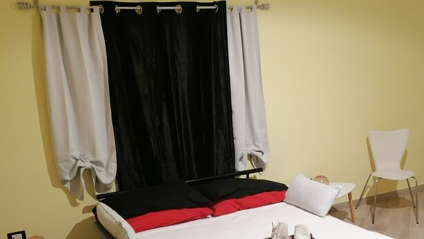 A neatly made bed with a white mattress, black and red pillows, and a white headboard. There is a small bedside table with a lamp, a chair, and a decorative item on the floor.