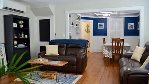 Una sala de estar con un sofá de cuero marrón, una mesa de centro de madera y una mesa de comedor con sillas. Hay una estantería y una planta en la habitación.