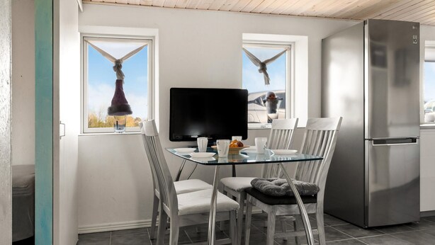 A modern kitchen with a glass dining table, white chairs, a stainless steel refrigerator, and a large window with curtains.