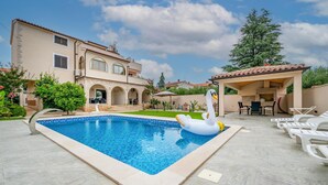 A swimming pool with a swan float, a covered patio with outdoor furniture, and a large house with a tiled roof. - Beautiful apartment in Medulin with swimming pool (Medulin)