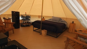 Un hébergement sous tente comprenant un lit, une table, une chaise, une étagère avec des vêtements pliés et un petit radiateur.