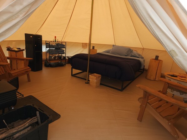 A tented accommodation with a bed, a table, a chair, a shelf with folded clothes, and a small heater.