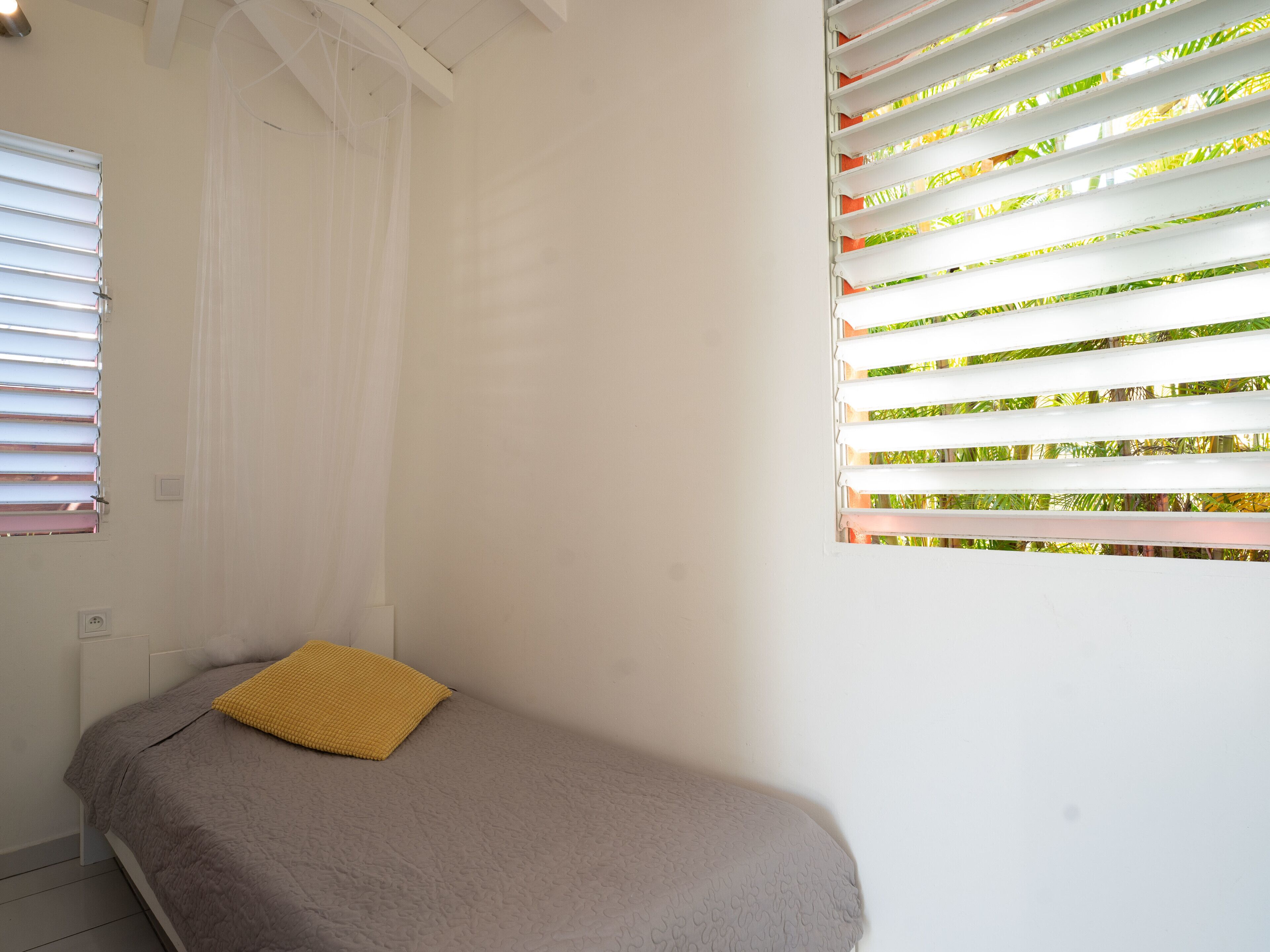 A small, single-bed room with a plain white wall, a window with white blinds, and a yellow pillow on the bed.