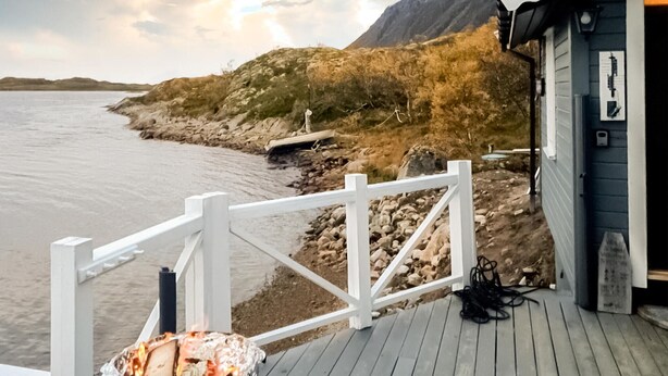 A wooden deck with a white railing, a small building with a dark exterior, and a rocky shoreline with a mountain in the background. - Awesome home in Laukvik with kitchen (Laukvik)