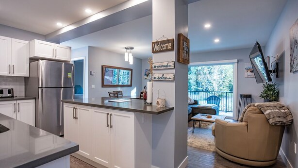 A modern kitchen with white cabinets, stainless steel appliances, and a central island. There is a dining area with a sofa and a TV mounted on the wall. - Al's Escape by Revelstoke Vacations (Revelstoke)