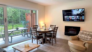 A dining area with a table set for four, a sofa, a TV mounted on the wall, and a balcony with a view of trees. - Al's Escape by Revelstoke Vacations (Revelstoke)