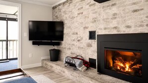 Una sala de estar con chimenea de ladrillo, un televisor de pantalla plana montado en la pared y una zona de estar acogedora con alfombra y manta.