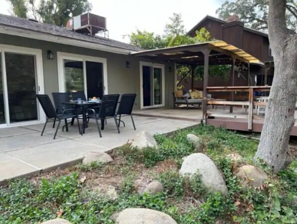 Enjoy dining or socializing in this outdoor space with a table and chairs, surrounded by lush plants and a pergola.