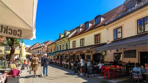 Une rue animée, bordée de cafés et de restaurants avec des terrasses, un ciel d’un bleu éclatant et une architecture européenne traditionnelle.