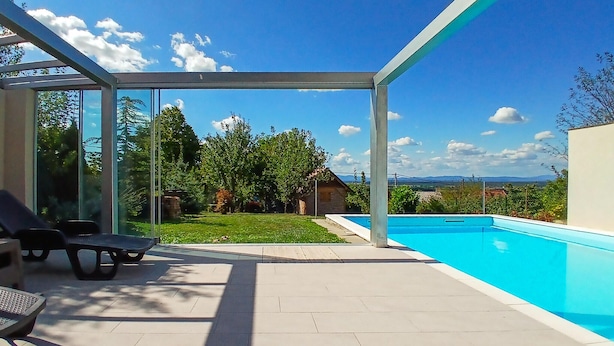 Un espace piscine extérieur moderne, avec une eau d’un bleu cristallin, entouré d’une structure en verre, et offrant une vue sur un paysage verdoyant et luxuriant, avec des arbres et une chaîne de montagnes lointaine.