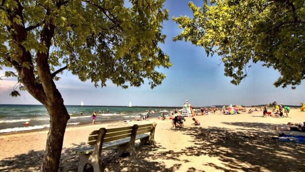A beach with a wooden bench, people sunbathing, and a lifeguard tower in the distance.