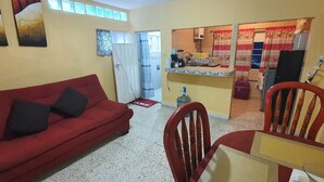 Una sala de estar con un sofá rojo, sillas de madera, una mesita y una zona de cocina con refrigerador y microondas.