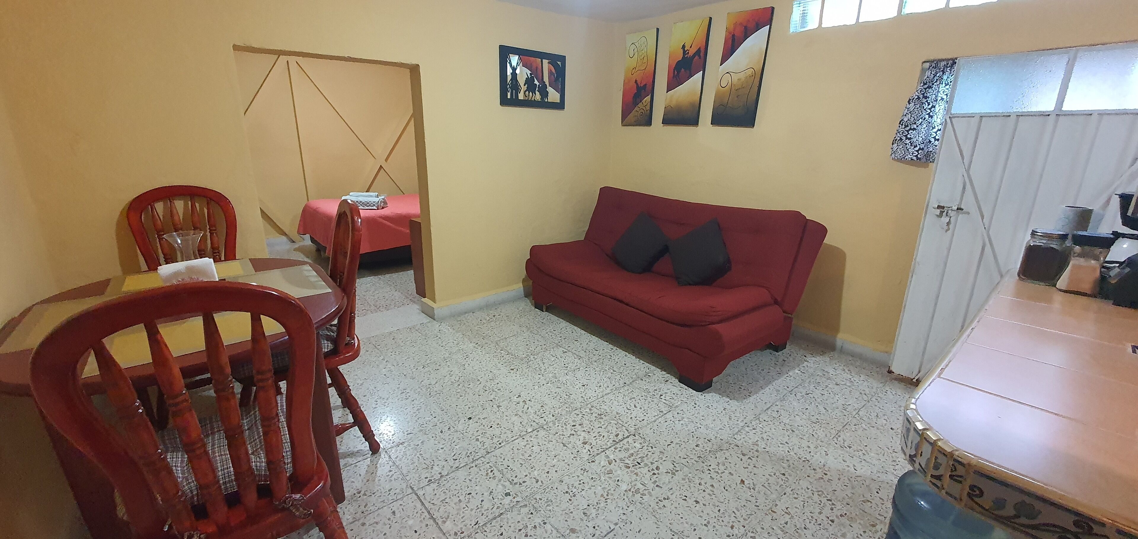 Una sala de estar con un sofá rojo, una mesa de comedor de madera con sillas, una cama pequeña con colcha rosa y una puerta que da a una zona de cocina.