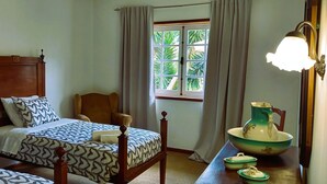 A bedroom with a wooden bed, a chair, a window with curtains, a lamp, and a table with a pitcher and plates. - Moradia Entre o mar e a ria (Gafanha da Nazaré)
