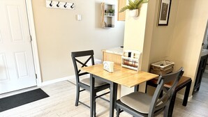 A dining area with a wooden table and four chairs, a small white shelf with books and a plant, and a door with a black doormat. - Historic District, Downtown, STE 3 (Rapid City)