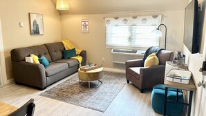 A living room with a sofa, armchair, coffee table, and ottoman. There is a painting on the wall, a window with blinds, and an air conditioning unit. - Historic District, Downtown, STE 3 (Rapid City)