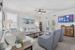 A living room with a sofa, armchairs, a coffee table, a ceiling fan, a TV, and a painting of boats.