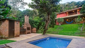 A swimming pool with a wooden deck, surrounded by lush greenery and a brick building. - Casa na Serra de Mulungu por Carpediem (Mulungu)
