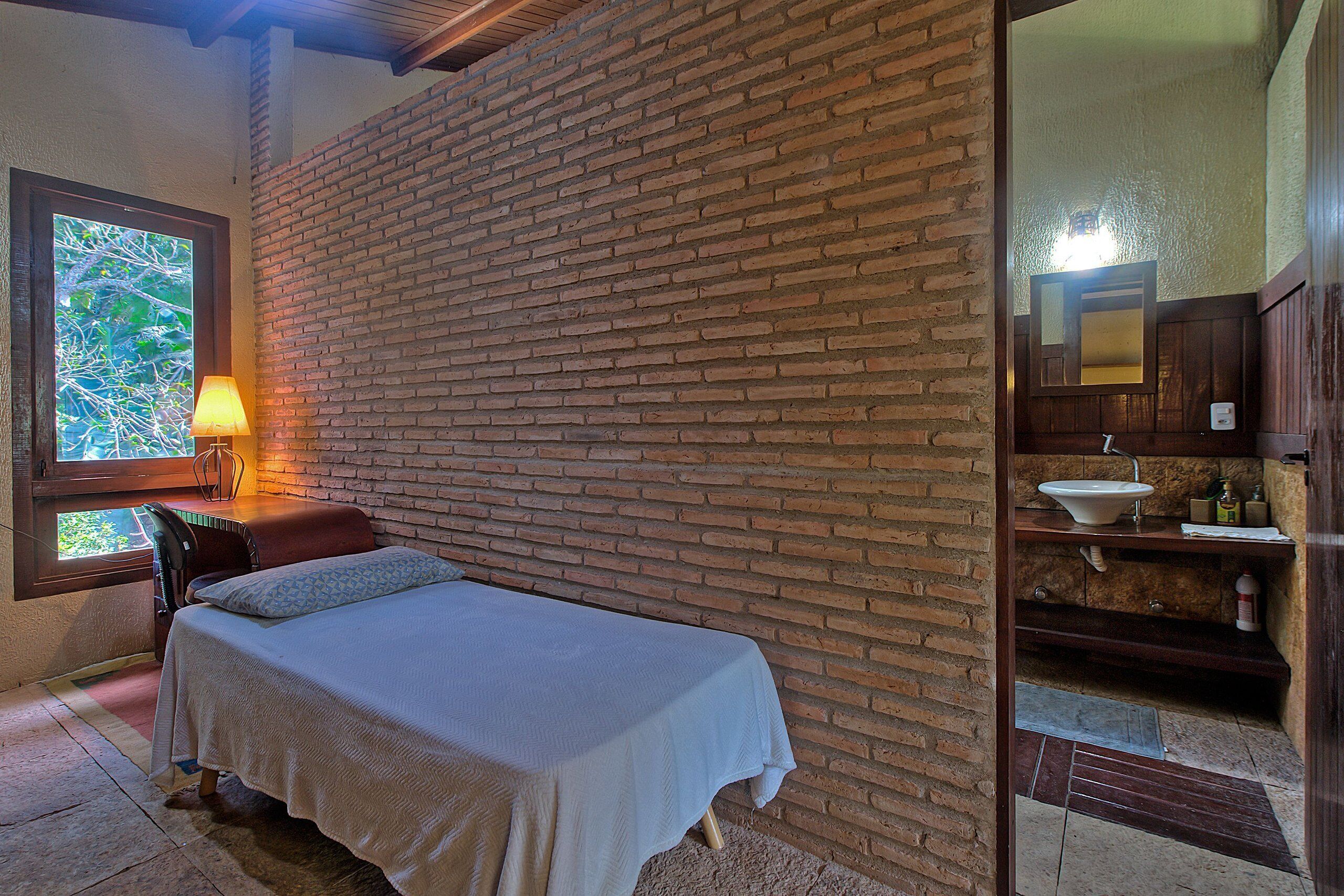 Una habitación con pared de ladrillo, una cama con cubrecama blanco, una ventana con vistas a árboles y un baño con lavamanos y espejo.