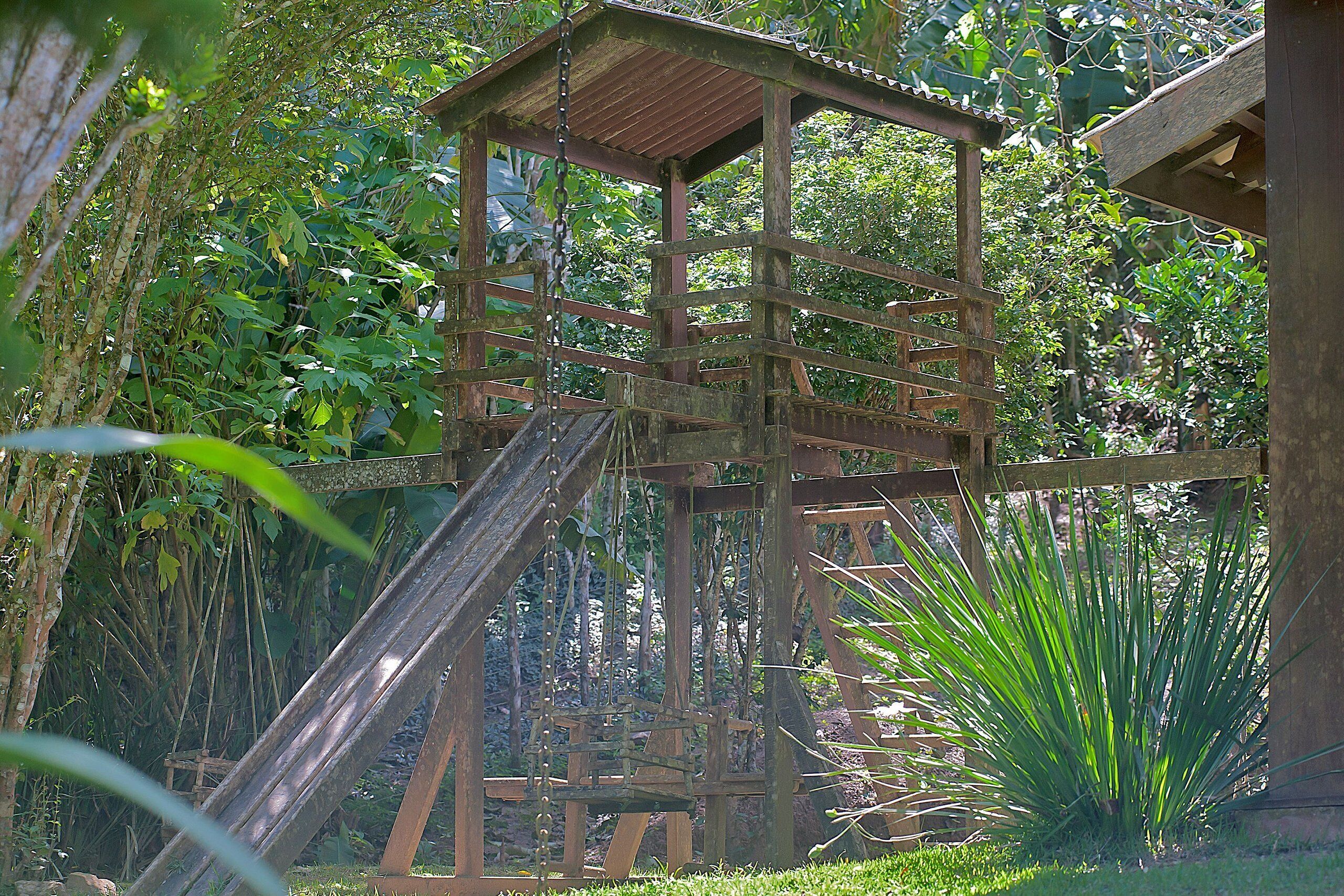 A wooden play structure with a slide, surrounded by lush greenery.