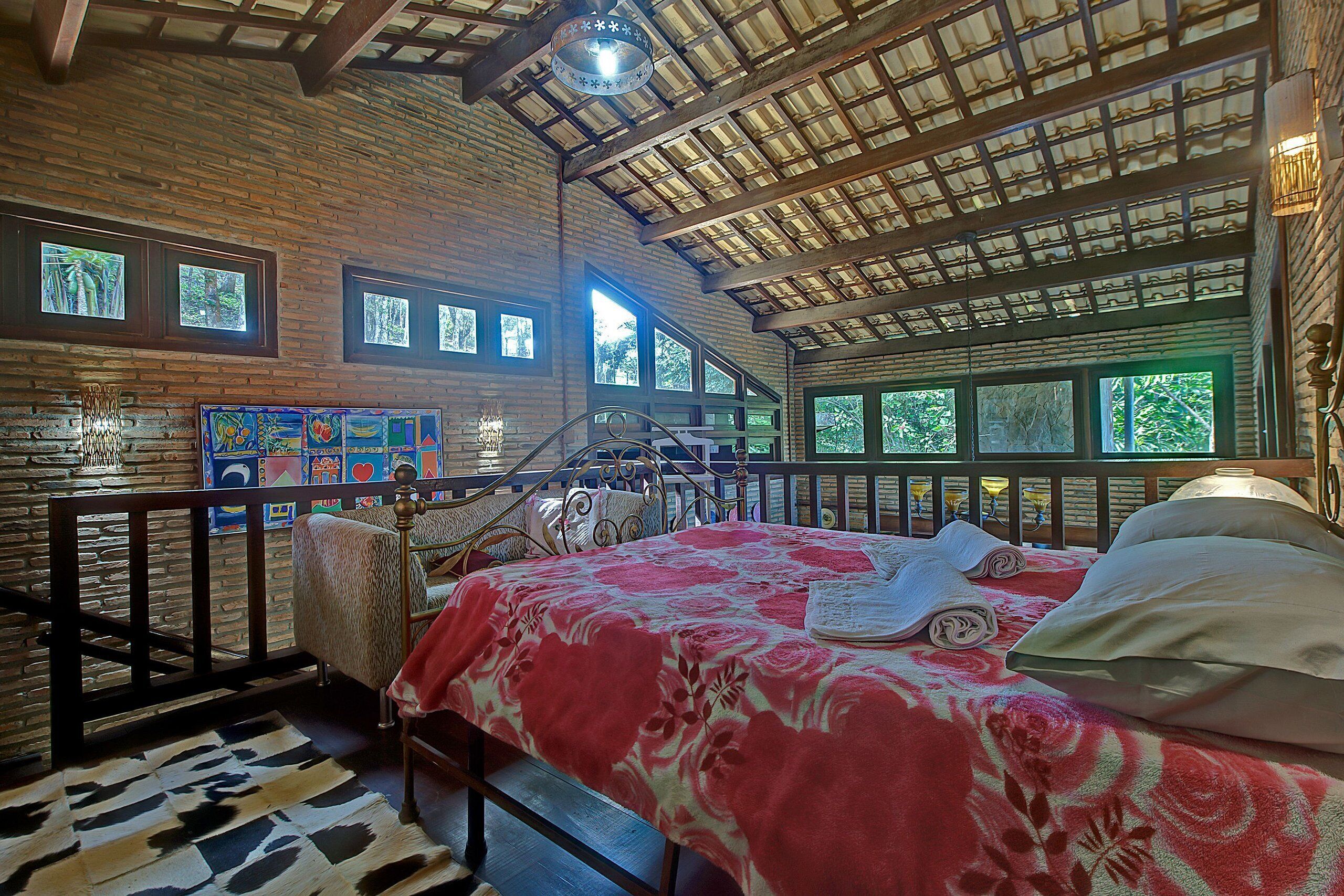 A spacious room with a large bed, a wooden ceiling, and multiple windows offering views of the outdoors.