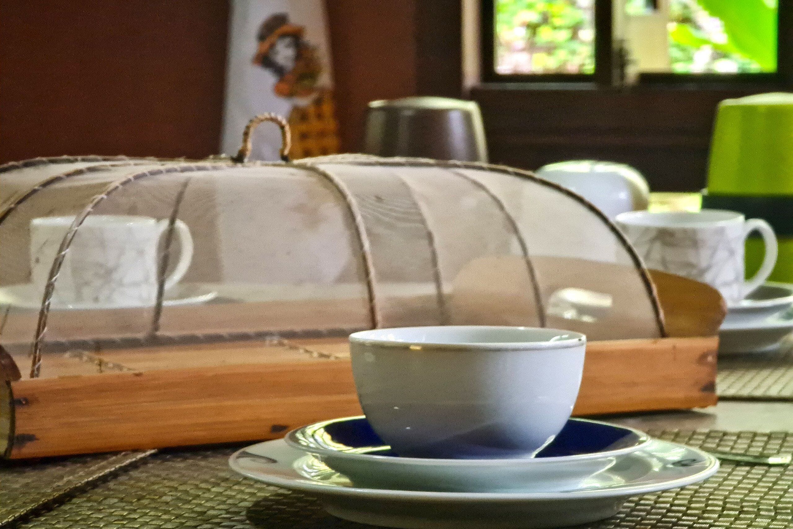 A table with a cup and saucer, a covered basket, and a green chair in the background.