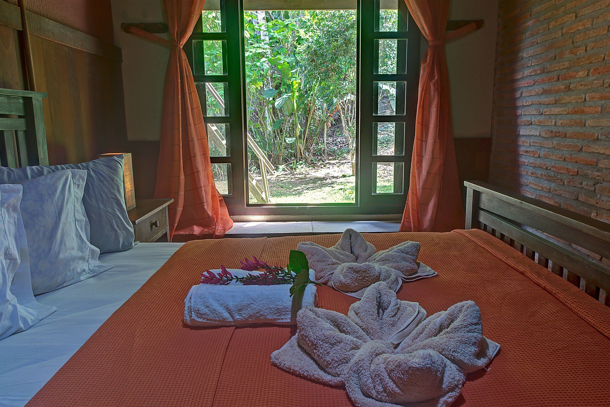 A hotel room with a bed, a window with curtains, and a view of greenery outside.