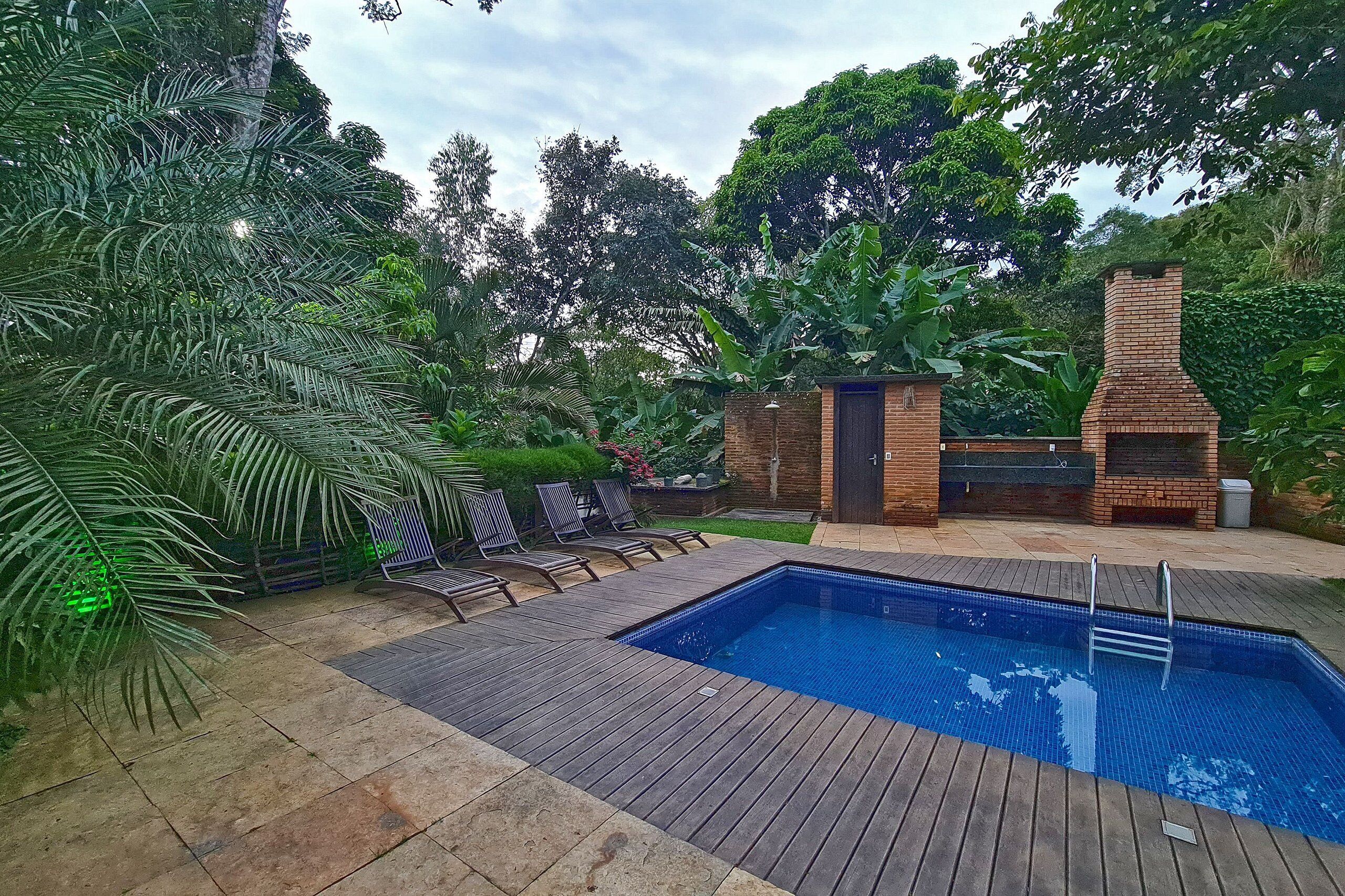 Un patio trasero con piscina, terraza de madera y zona de barbacoa de ladrillo rodeada de abundante vegetación.