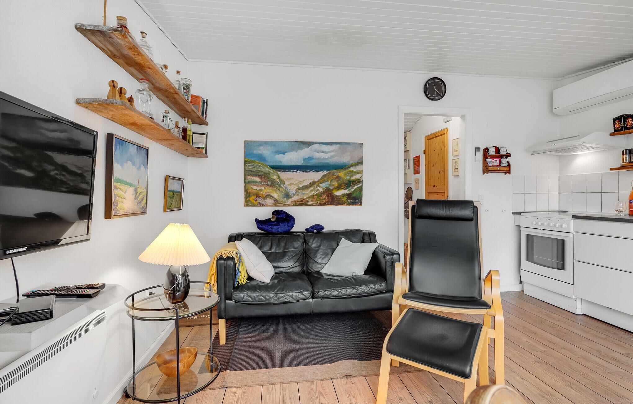 Un salon avec un canapé en cuir noir, une table basse en verre, un tableau au mur et un coin cuisine doté de meubles blancs et d’une cuisinière.