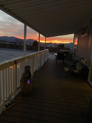 A covered deck with a railing, a table, and chairs, overlooking a snowy landscape with mountains in the distance.
