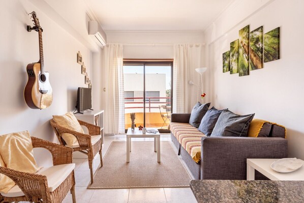 A living room with a sofa, two armchairs, a coffee table, and a guitar. There is a balcony with a view of a building.