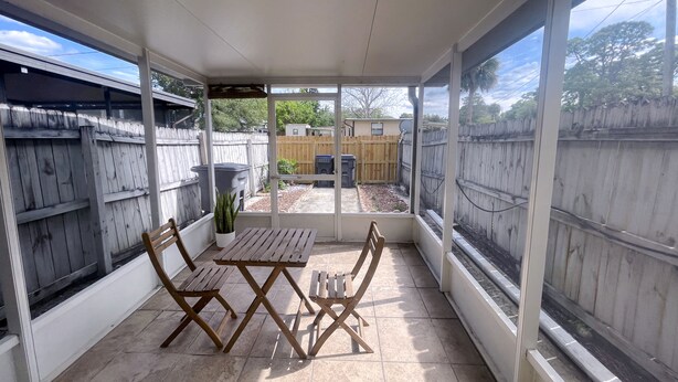 Un patio techado con una mesa y sillas de madera, una planta en maceta, y vistas a un jardín trasero con cerca de madera y una antena parabólica.