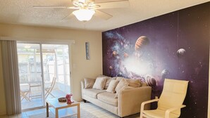 Una sala de estar con un sofá color beige, una mesa de centro de madera, un sillón blanco y un ventilador de techo. Se ve un patio trasero a través de una ventana grande.