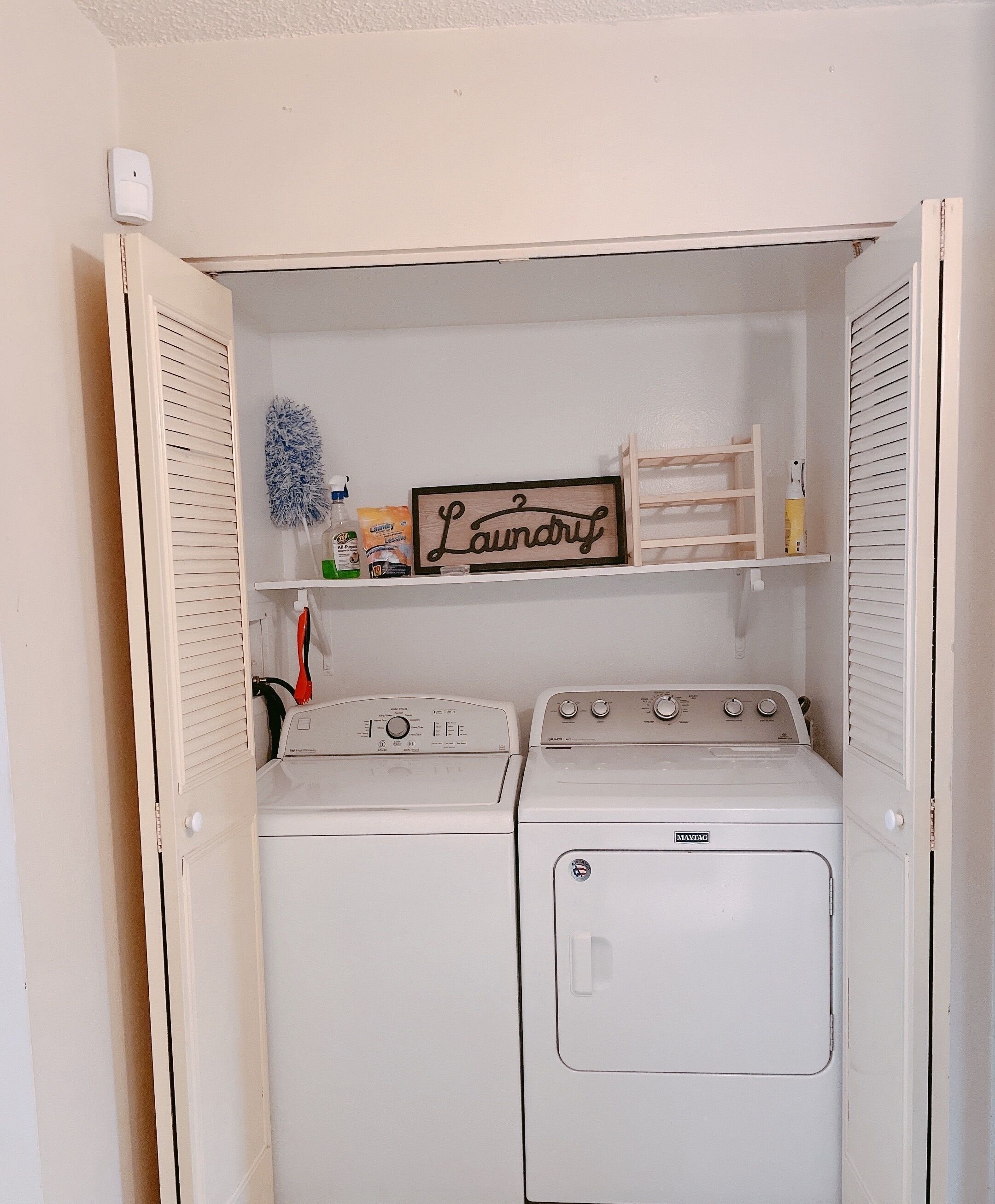 Ein Waschraum mit zwei übereinander gestapelten, frontbeladenen Waschmaschinen und Trocknern, einem Holzschild über den Trocknern und einem Regal mit Waschmitteln.