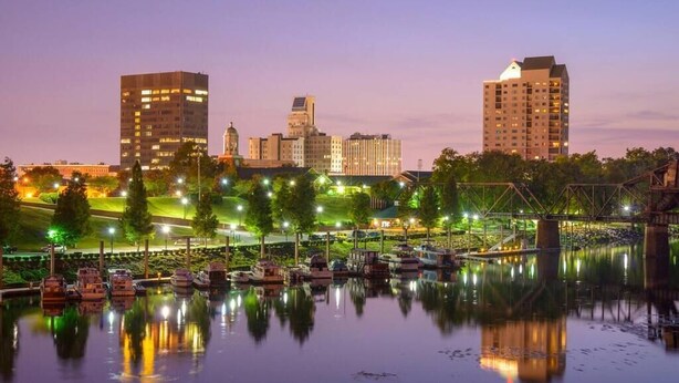 Un paisaje urbano frente al agua con un puente, barcos y edificios iluminados al atardecer.