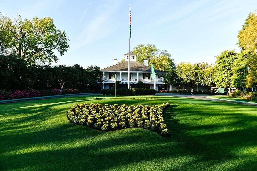 A well-manicured lawn with a distinctive flower bed in the shape of a state outline, a flagpole with a flag, and a large building with a porch.