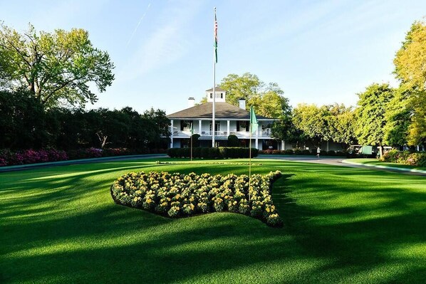 A well-manicured lawn with a distinctive flower bed in the shape of a state outline, a flagpole with a flag, and a large building with a porch.