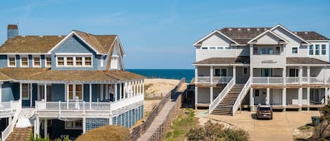 Una zona residencial costera con dos casas frente a la playa, una con balcón y la otra con porche y escaleras que llevan a la entrada.