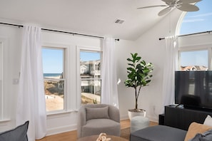 A modern living room with large windows, a ceiling fan, a flat-screen TV, and a potted plant. - LUX Oceanside, Pool, Hot Tub, Decks, Arcade, Pets, Bikes, Loaded (Nags Head)