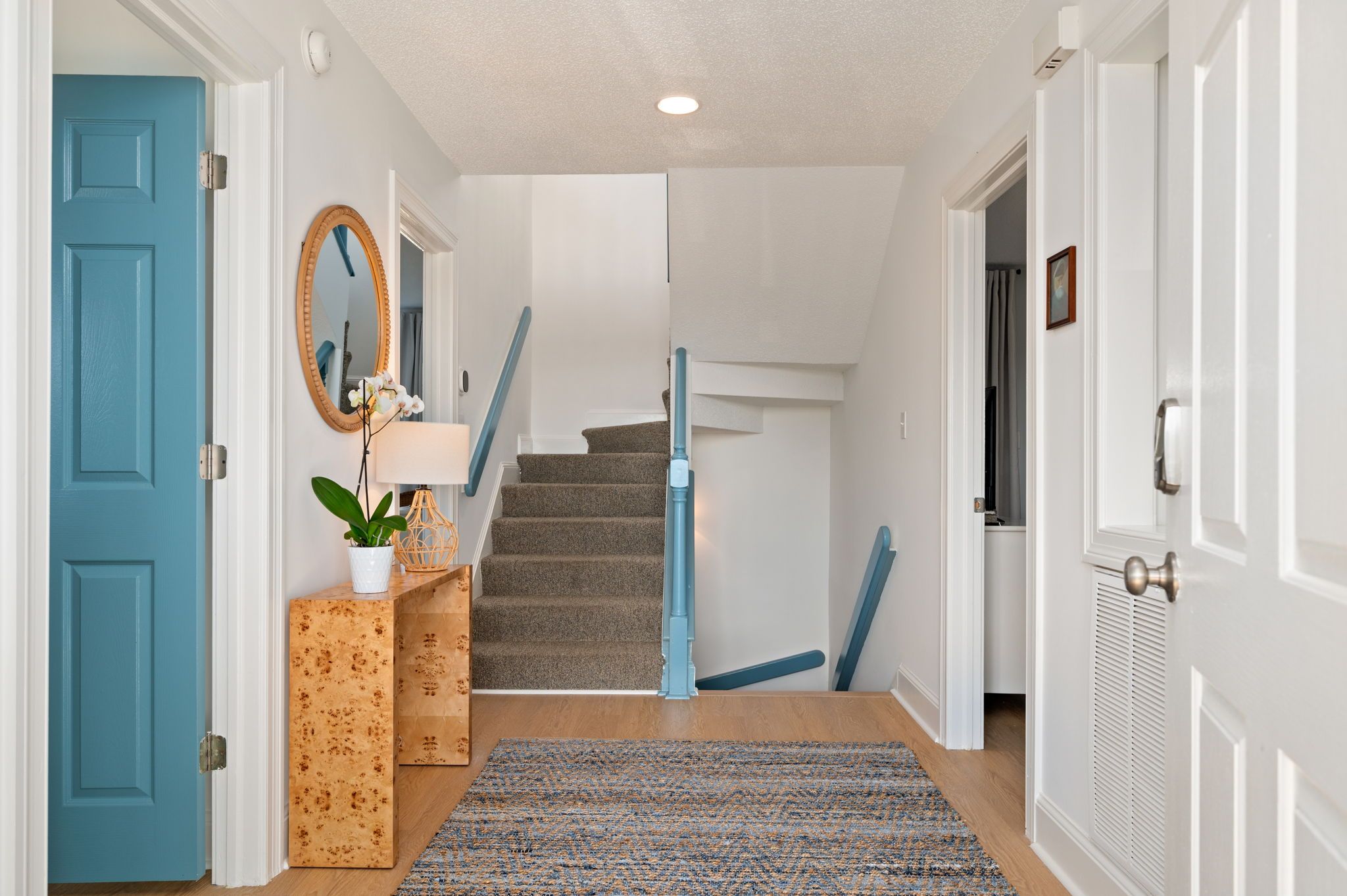 A hallway with a blue door, a mirror, a plant, a lamp, a staircase, and a patterned rug.