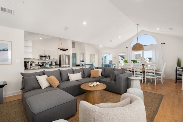 A modern living room with a sectional sofa, a round coffee table, and a dining area with a table and chairs. - LUX Oceanside, Pool, Hot Tub, Decks, Arcade, Pets, Bikes, Loaded (Nags Head)