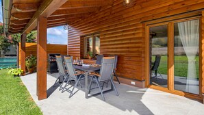 Une terrasse extérieure avec une table et des chaises en bois, un barbecue et une vue sur une piscine et la verdure.