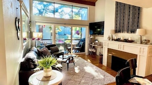 A living room with a sofa, coffee table, and fireplace. There is a large window providing natural light and a view of the outside.