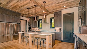 A modern kitchen with a wooden ceiling, stainless steel appliances, and a central island with stools. - Manna Mountain Lodge ~  Indoor Pool, XBox Room, Gaming Garage, Views & More! (Sevierville)